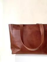 Cartera tipo tote de cuero graneado color negro, con textura uniforme y suave al tacto. Tiene manijas de 55 cm, bolsillo frontal con broche imantado, bolsillo trasero con cierre metálico, cierre principal metálico, forro interior y tres bolsillos internos (uno con cierre, uno plaqué y uno tipo sobre con solapa y botón).