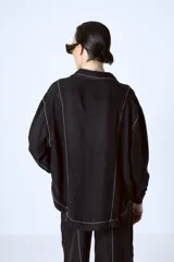 Blusa negra con botones, cuello camisero y mangas largas con puños. Presenta pespuntes blancos en contraste.