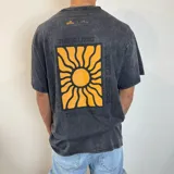 Remera negra con efecto gastado, corte oversize y estampado en la espalda con un sol amarillo dentro de un rectángulo, con las frases "This is living" y "Sunsets Sessions".