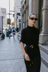 Vestido negro de tejido de punto, corte midi y silueta holgada, con cuello alto acanalado y mangas cortas tipo murciélago. Se ajusta a la cintura con un cinturón fino de efecto piel negro con hebilla dorada en forma de C.