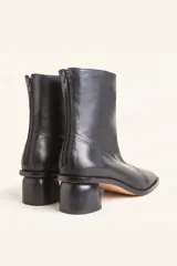 Bota corta de cuero vacuno color marrón oscuro, con diseño sin forro y taco cilíndrico de madera tallada a mano. Presenta cierre posterior y punta redondeada.