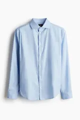 Camisa celeste de algodón con cuello italiano, tapeta clásica y canesú en la espalda. Mangas largas con tapeta y botón, y cierre ajustable de botón en los puños. Bajo levemente redondeado. Corte ajustado que realza el contorno del cuerpo creando una silueta entallada.