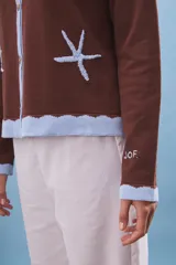 Cárdigan marrón de tejido de punto con cuello polo celeste, cierre frontal con botones y dos estrellas de mar bordadas.