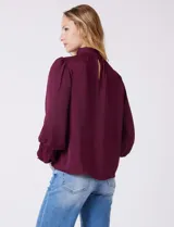 Blusa de gasa satén color bordó, marca Tahari, con cuello alto fruncido y mangas largas abullonadas. La espalda tiene una abertura central con botón.