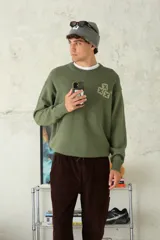 Sweater de tejido de punto color verde oliva, con cuello redondo y puños y ruedo acanalados. Presenta un diseño bordado de dos cruces superpuestas en el pecho.