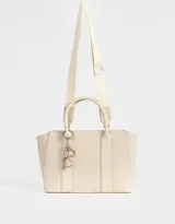 Cartera tipo tote color blanco crudo, con doble asa corta y correa larga regulable y desmontable. Incluye charm colgante con forma de corazón y cierre superior.