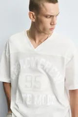 Camiseta blanca de corte holgado con cuello en V y mangas cortas. Presenta un diseño de malla con diferentes texturas combinadas a tono y estampado frontal con la inscripción "Legacy Crew 95 Elite Mode".