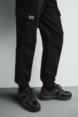 Pantalón cargo negro de corte slim fit, confeccionado en tejido de algodón con cintura elástica ajustable con cordón. Cuenta con bolsillos frontales, traseros y bolsillos tipo parche con solapa en los laterales.