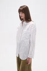 Camisa de algodón blanca de corte holgado, con cuello camisa y manga larga. Presenta un estampado de finas rayas verticales negras y un bolsillo frontal con rayas horizontales.