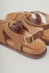 Sandalia plana de gamuza color marrón oscuro, con tiras cruzadas en la parte delantera y una tira fina ajustable con hebilla metálica en el tobillo. Posee plantilla de cuero y suela baja.