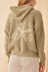 Campera color verde militar de punto inglés con capucha y terminaciones rotas en los puños. Tiene un bordado artesanal en la espalda con motivos marinos.