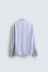 Camisa celeste a rayas verticales blancas, de corte recto, con cuello italiano abierto, mangas largas con puños abotonados y cierre frontal con botones.