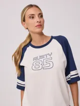 Remera bicolor de corte holgado, con cuerpo blanco y mangas ranglán azul marino con dos rayas blancas en los puños. Estampado frontal con la palabra "Rusty" y el número "85" en azul.