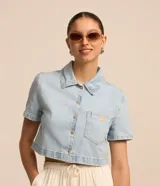 Camisa cropped de jeans celeste con cuello clásico, mangas cortas, cierre frontal con botones y bolsillo en el pecho.