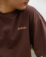 Remera marrón de corte oversize confeccionada en jersey de algodón, con estampa en la espalda con el texto "The Tribu Brand Creative Studio" en color crema y un diseño de tres líneas horizontales debajo.