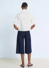 Camisa blanca de popelín de algodón, manga tres cuartos, corte crop y diseño recto. Presenta cuello camisero, bolsillo en el pecho con detalle de strass y cierre frontal con botones.