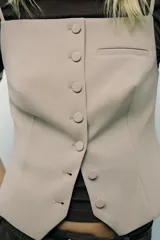 Chaleco color beige de escote recto y tirantes finos. Presenta un falso bolsillo de vivo en el delantero y cierre frontal con botones forrados.