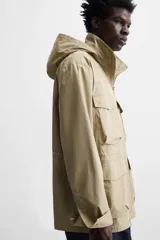 Parka color beige de tejido técnico ligero, corte relaxed fit, cuello alto con capucha plegable, manga larga con puños ajustables con botón a presión, cierre frontal con cremallera, cintura ajustable con elástico interno y múltiples bolsillos frontales de parche con solapa.