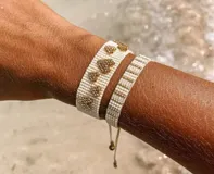 Set de dos pulseras tejidas con cuentas. Una pulsera es de color blanco con corazones dorados, y la otra es de color blanco con rayas doradas.