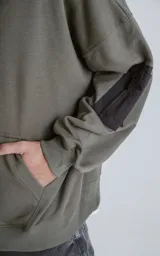 Hoodie color verde militar de rústico 100% algodón, con silueta oversize, capucha forrada y bolsillo tipo bomber en la manga con cierre. Presenta bolsillo canguro frontal.