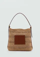 Bolso tipo shopper de paja trenzada color beige con aplique de cuero marrón en el frente. Tiene cierre con botón magnético, bolsillo interior, detalles calados y asa de hombro ajustable con hebillas.
