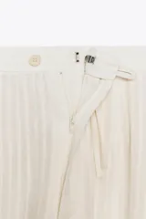 Pantalón de tiro medio color crudo con rayas verticales, confeccionado en hilatura con lino. Tiene bolsillos laterales ocultos en costura y de vivo en espalda, detalle de pliegues y bajo acabado en puño.