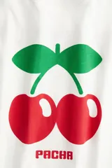 Remera blanca de algodón de corte amplio con cuello redondo y mangas cortas. Estampado frontal de dos cerezas rojas con hojas verdes y el logo de la marca Pacha en rojo.