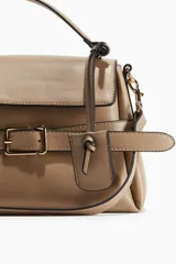 Cartera bandolera color visón, con correa ajustable y asa de mano. Presenta solapa con cierre magnético y trabilla decorativa con hebilla metálica.