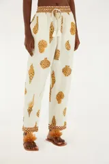 Pantalón de corte recto con estampado de conchas marinas en tonos marrones sobre fondo blanco. Presenta cintura elástica ajustable con cordón y borlas, bolsillos laterales y dobladillo con detalle de ribete marrón.