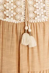 Vestido corto color beige con cuello redondo, cordón con borlas y manga larga. Presenta bordados combinados a contraste en la pechera, cierre frontal con botones ocultos, forro interior y bajo con volantes.