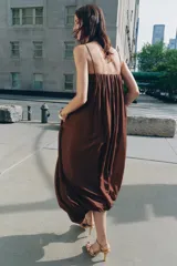 Vestido largo marrón de tejido satinado, con escote recto, tirantes finos extraíbles, bolsillos laterales ocultos y bajo asimétrico acabado en globo.
