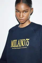 Sudadera de tejido de punto color azul marino, con cuello redondo y mangas raglán. Presenta un estampado frontal en amarillo con el texto "MILANO 75" y una dirección debajo.