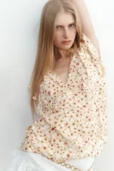 Blusa de tejido crepé color crema con estampado de flores pequeñas en tonos rosados y marrones. Tiene corte holgado, cuello con volados y escote en V con tiras para ajustar. Las mangas son cortas tipo mariposa con ribete de volado.