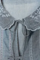 Camisa corta celeste confeccionada en algodón 100%. Presenta cuello bobo con bordados florales, mangas cortas abullonadas y cierre frontal con lazadas.