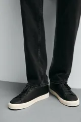 Zapatilla urbana de cuero sintético color negro, con cordones y suela de goma color crema.