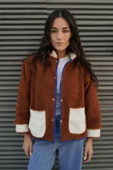 Campera marrón de tela tipo sherpa con cuello, puños y bolsillos de tela blanca. Tiene cierre con botones a presión y ribete azul.