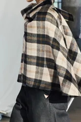 Capa de cuello alto confeccionada en tejido con lana a cuadros en tonos beige, negro y marrón. Tiene bajo asimétrico y cierre en la espalda con botón.