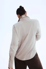 Blusa de manga larga color gris claro o beige pálido, con acabado satinado y cuello clásico que forma un escote en V profundo con cierre de botones frontales.