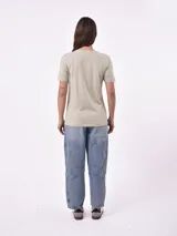 Remera blanca de algodón con cuello redondo y manga corta. Presenta un pequeño bordado en el pecho izquierdo con una figura que parece ser una persona acostada en una tabla de surf o flotador.