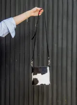 Cartera tipo canasta de cuero con estampado animal print de vaca en blanco y negro. Tiene correa larga de cuero negra y herrajes metálicos plateados.