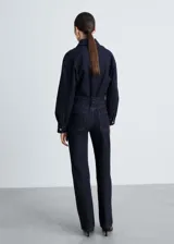 Mono largo de denim azul oscuro, con cuello camisero, mangas largas con puños abotonados, trabillas, cinturón con hebilla metálica, cierre frontal con botones a presión y cuatro bolsillos.