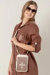 Bolso pequeño tipo bandolera o cartera de mano, confeccionado en cuero vacuno color taupe/marrón claro, con detalles en material metalizado plateado. Presenta un bordado de flor de lis en el frente y cierre superior. Incluye correa fina desmontable.