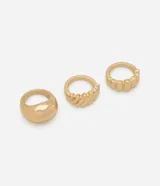 Set de tres anillos dorados de metal con diferentes diseños: uno liso y grueso, otro con textura de líneas diagonales y otro con textura de bloques rectangulares.