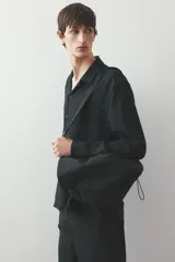 Camisa negra de tejido suave con cuello abierto, tapeta lisa y canesú en la espalda. Modelo de manga larga con cierre ajustable de botones en los puños y tapeta con botón. Bajo recto y corte estándar.