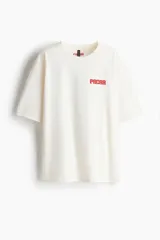 Remera blanca de corte amplio con cuello redondo y mangas cortas. Estampado pequeño con la palabra "PACHA" en rojo en el lado izquierdo del pecho.