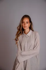 Camisa color beige claro, de corte holgado, con cuello clásico, cierre frontal con botones, mangas largas con puños abotonados y detalles de broderie en la parte delantera.