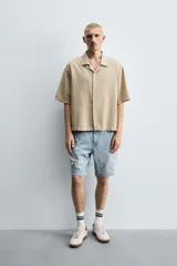 Camisa de denim de algodón, color beige claro, con corte relajado, cuello bowling y manga corta. Presenta cierre frontal con botones a presión y un pequeño bordado a tono en el pecho.