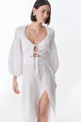 Vestido color beige de lino, con escote en V, mangas largas abullonadas y lazos frontales. Tiene corte a la cintura y falda larga con tajo frontal.