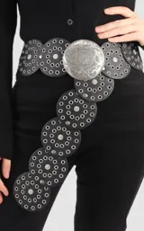 Cinto negro con apliques circulares de cuero y tachas plateadas. Hebilla circular plateada con diseño grabado.