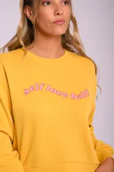 Buzo amarillo con cuello redondo y estampado frontal con la frase 'self love baby' en color rosa.